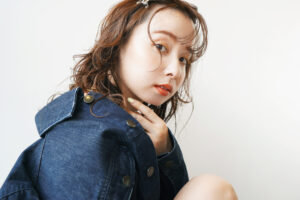 ゆるいパーマ風ウェーブのミディアムヘアを、顔まわりに沿う束感と透け感バングで軽やかに見せ、デニムジャケットに合わせてこなれた雰囲気に仕上げたサロンスタイル写真。
