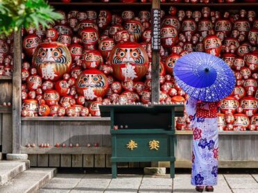 大阪・箕面の勝尾寺で、奉納棚いっぱいに並ぶ赤いだるまの前に、浴衣姿の人物が青い和傘を差して佇む風景写真。